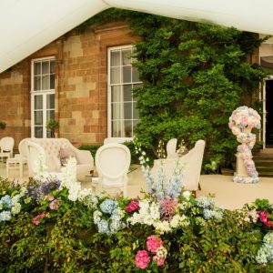 Photo shows area where Dining Room Terrace Marquee connects to the side of Hillsborough Castle. Cream carpets cover the stone terrace and comfortable armchairs and planting added to create an outside-in-feel.