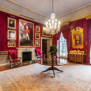View of Red Room - walls adorned with silk red damask. Walls covered with gold-framed paintings. A Waterford Crystal chandelier hangs above a small round mahogany table.