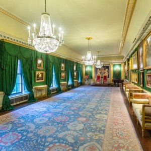 Throne Room with silk green damask walls, wooden floor and large rug. Two red and gold Chairs of State, or 'thrones' are situated at the far end of the room. Gold-framed paintings of landscapes adorn the walls and three large chandeliers hang above.